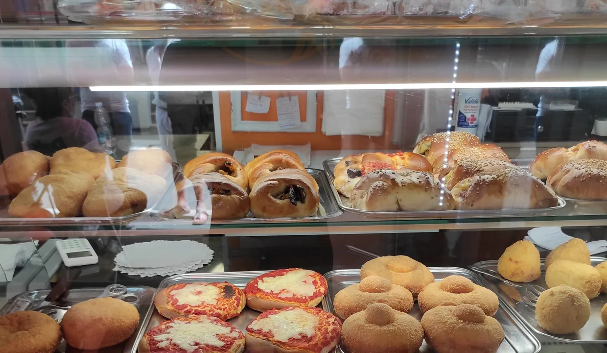 Il banco visto dal vetro: teglie di pizzette, bocconcini al sesamo, arancine e vaschette di pasta al forno.